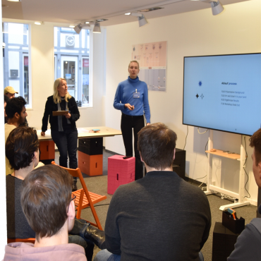 Mehrere Schüler*innen bekommen einen Vortrag. Vor ihnen erklärt eine blonde Frau in einem blauen Pullover etwas. Sie steht an einem Monitor.